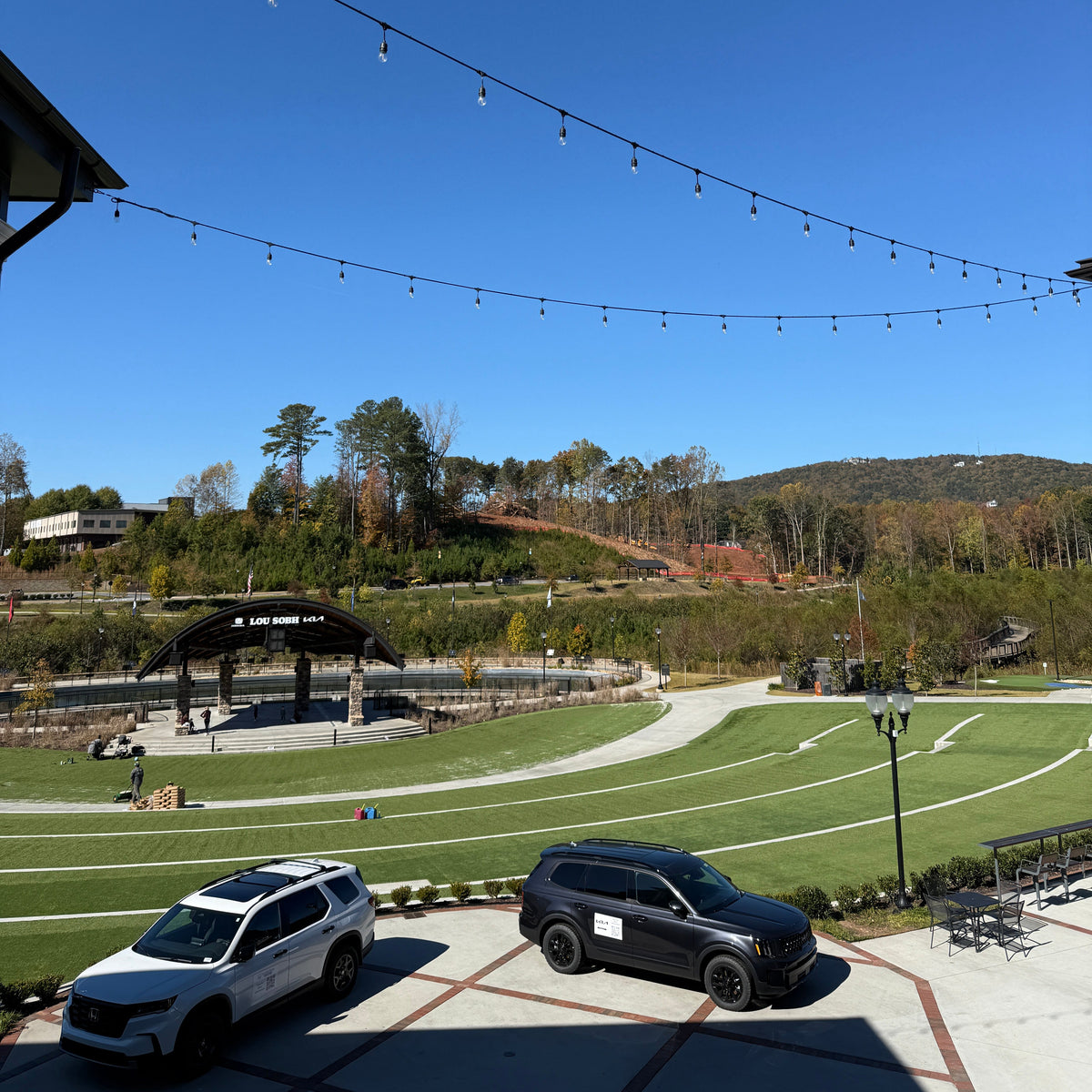 An outdoor landscape view of a sports field with two SUVs parked in the foreground, a covered pavilion, trees, and hills under a clear blue sky. String lights hang across the top of the frame.