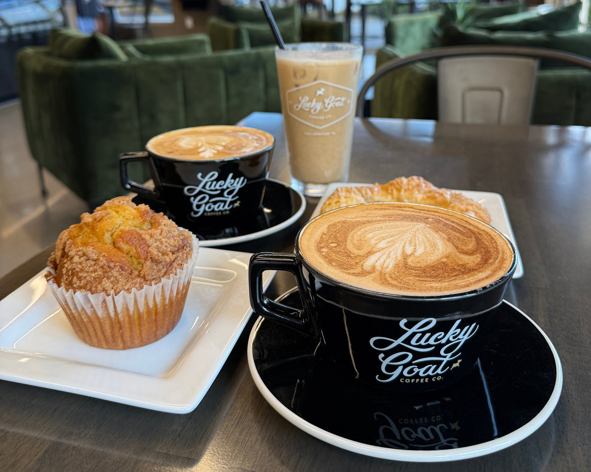 A cozy cafe table scene featuring a black ceramic cup of latte art coffee with 'Lucky Goat Coffee' branding, a matching cup of cappuccino, an iced coffee, and a muffin on a white plate, set against a blurred cafe background.