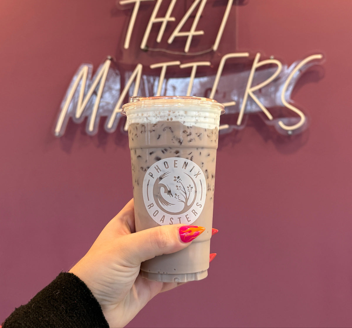 A hand with orange nail polish holds a cup of Phoenix Roasters iced coffee in front of a pink wall with a glowing 'COFFEE THAT MATTERS' neon sign.