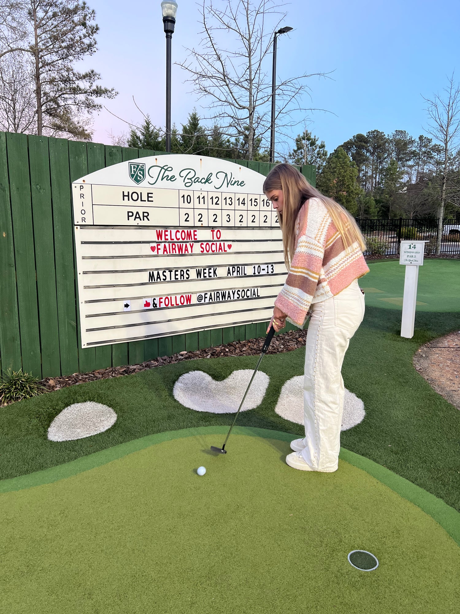A woman in a pink and white striped sweater and white pants is playing golf on a mini golf course, hitting a ball toward a hole on a green putting surface with a sign reading 'The Back Nine' in the background.