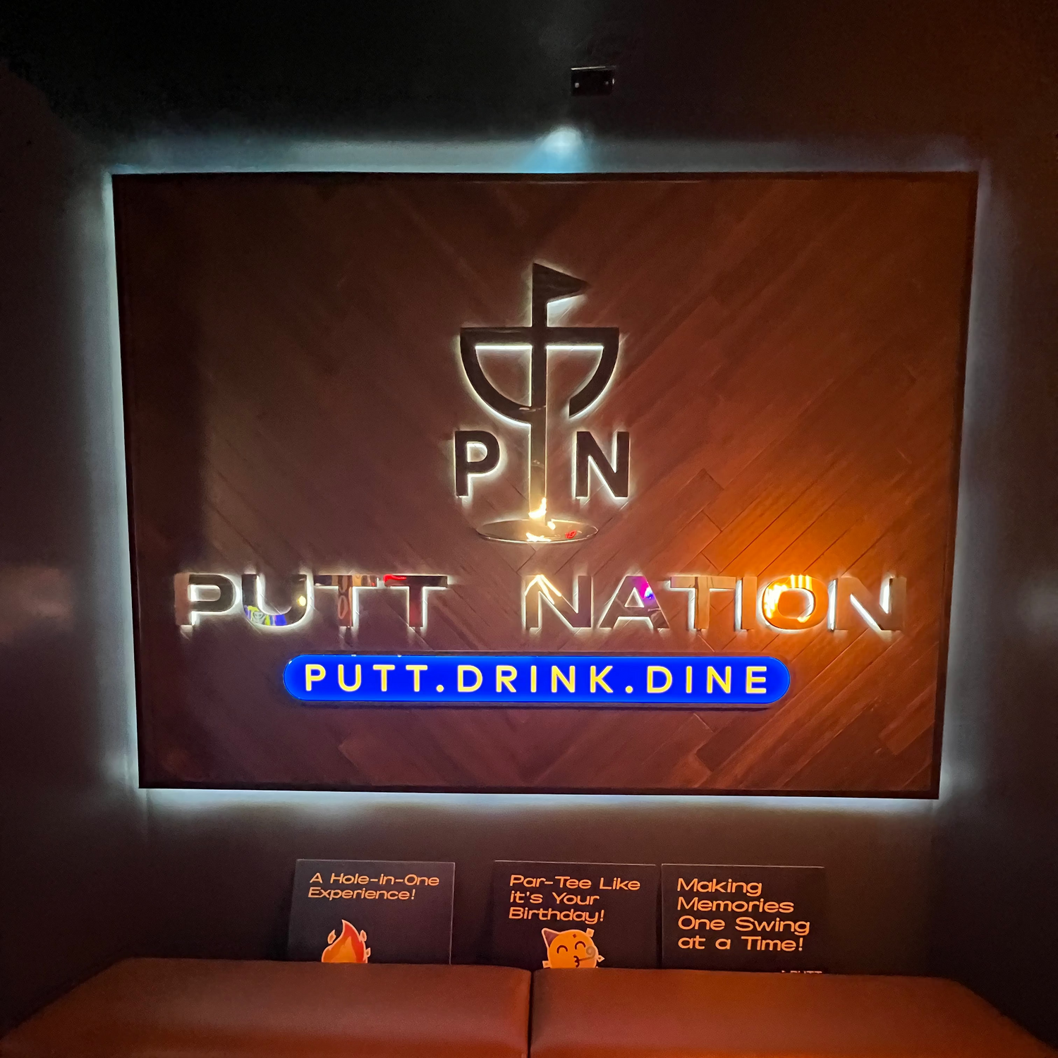 A dimly lit indoor space with a large illuminated wall sign reading "PUTT NATION" and "PUTT.DRINK.