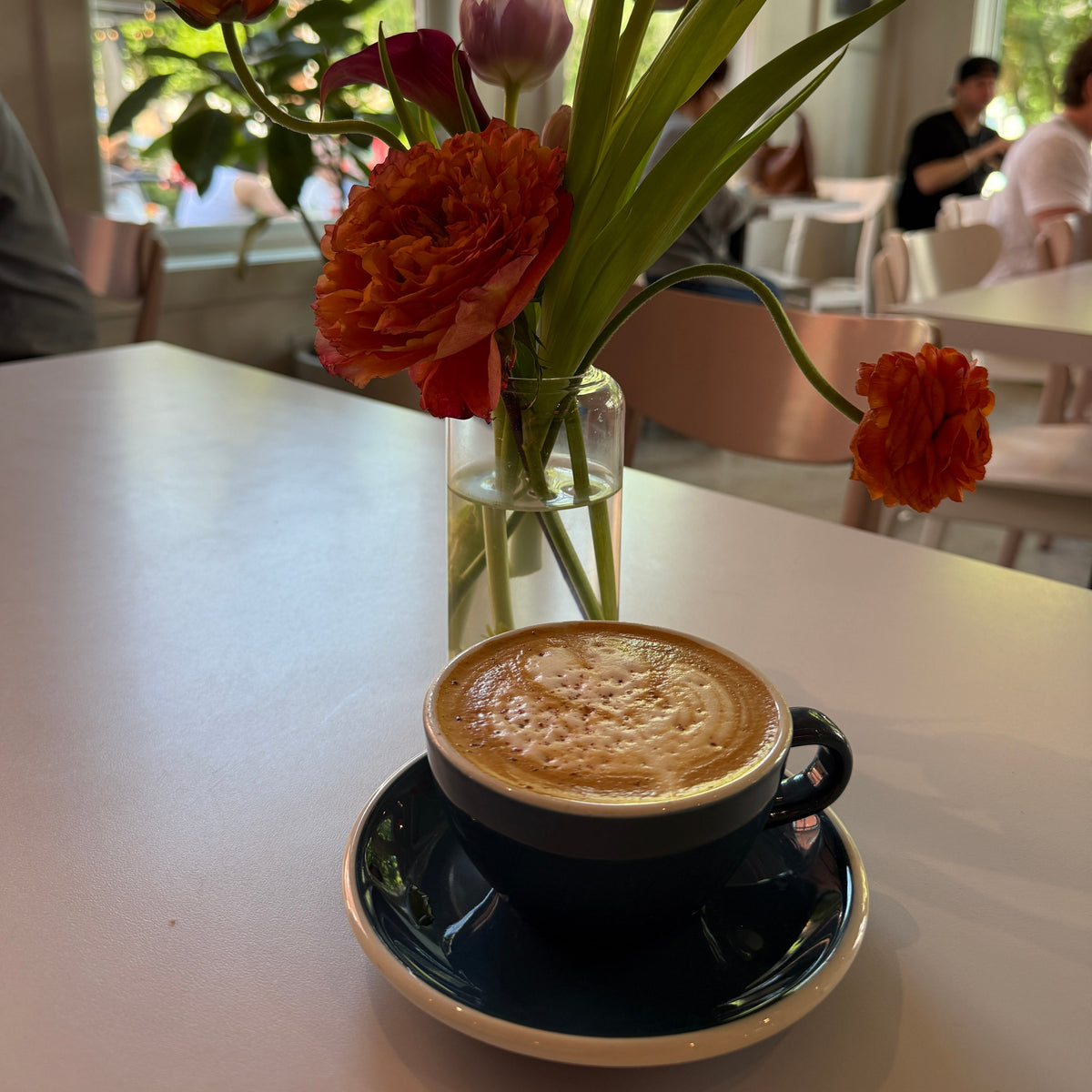 A cup of cappuccino on a saucer sits on a white table in a bright cafe, with a small glass vase of orange and pink flowers behind it.
