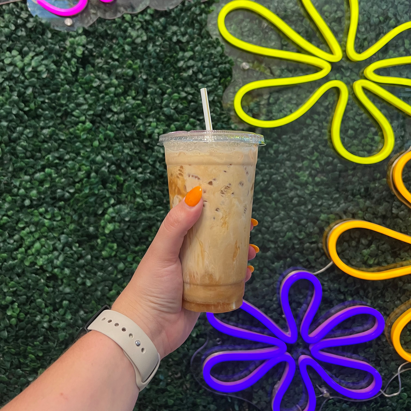 A hand holding a clear plastic cup of iced coffee with a white straw, set against a green artificial grass background with colorful neon flower and script signs.