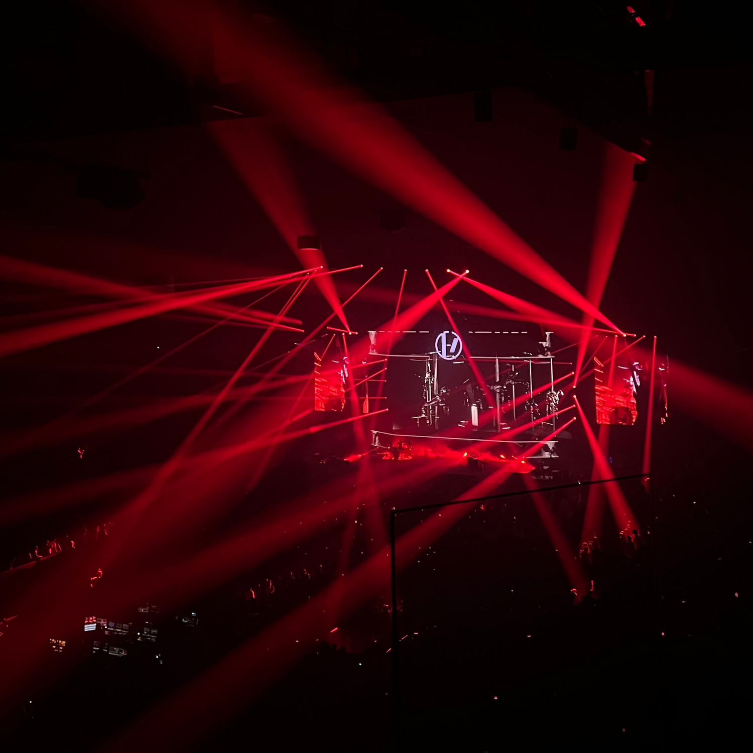A concert stage illuminated by dramatic red laser beams cutting through a dark arena, with a small lit performance area in the center.