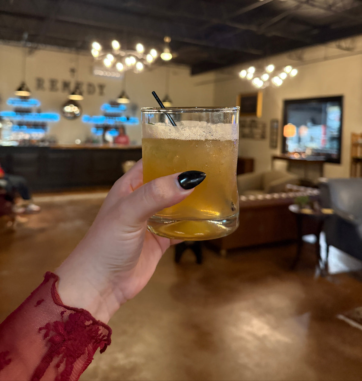 A hand with black nail polish holds a clear glass containing an amber-yellow beverage with a foamy top and a black straw, set against a blurred indoor bar or lounge environment with a bar counter and hanging lights in the background.