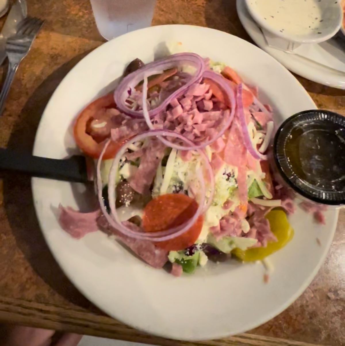 A close-up view of a prepared food dish on a restaurant table, featuring a white plate with sliced tomatoes, red onion, ham, and other toppings, alongside glasses of water and other dining items.